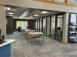 Area in library with tables, chairs, and enclosed study rooms with floor to ceiling windows and glass doors.